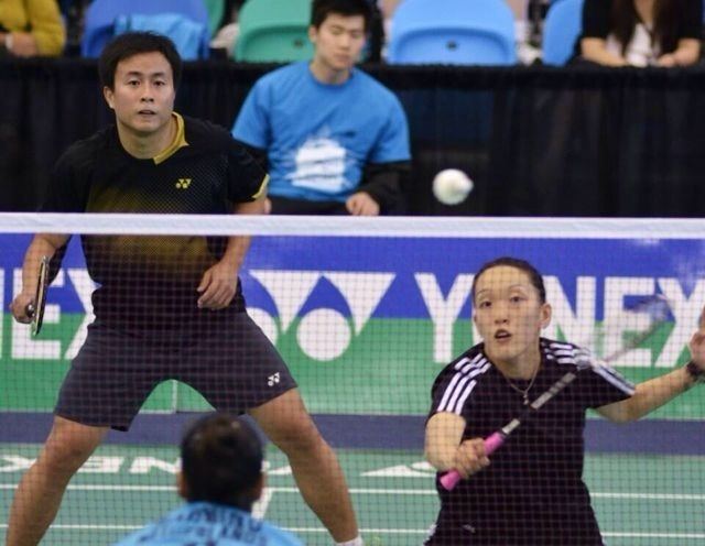 In a badminton court with an audience at the back with a blue chair, with a man sitting as line guides. From left, Kennevic Asuncion is serious, playing badminton, right hand holding a badminton racket, he has black hair wearing a black shirt and black shorts with white socks, at the right, Eva Lee is serious, holding a pink badminton racket she has black short-bald hair wearing a black shirt with white lines across the shoulder, a necklace and a black strap bracelet. In front is a badminton net, a division of two courts with an opposite team behind has black hair wearing a blue shirt.