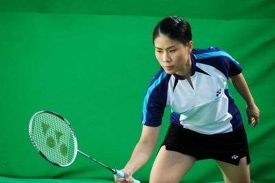 In a green room Kennie Asuncion is serious, posing, leaning with his right foot forward while holding a badminton racket. He has black hair and is wearing a white polo with blue sleeves and collar along with a black shorts and a white shoes with white socks.