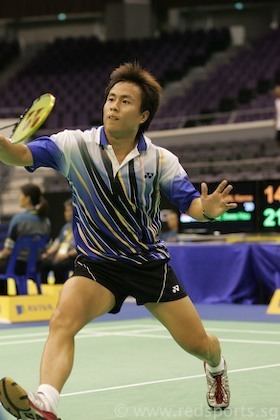 In a badminton court, with empty sits at the back and tables with blue cloth, Kennevic Asuncion is serious, leaping with his right foot forward, holding a badminton racket, he has black hair wearing a black strapped watch, white printed shirt with blue collars and sleeves, a black shorts along with a white shoes and socks.