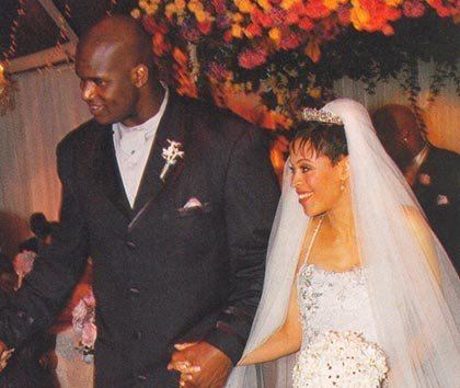 Kenny Anderson wearing a suit and his wife wearing a white gown holding a bouquet of white flowers.