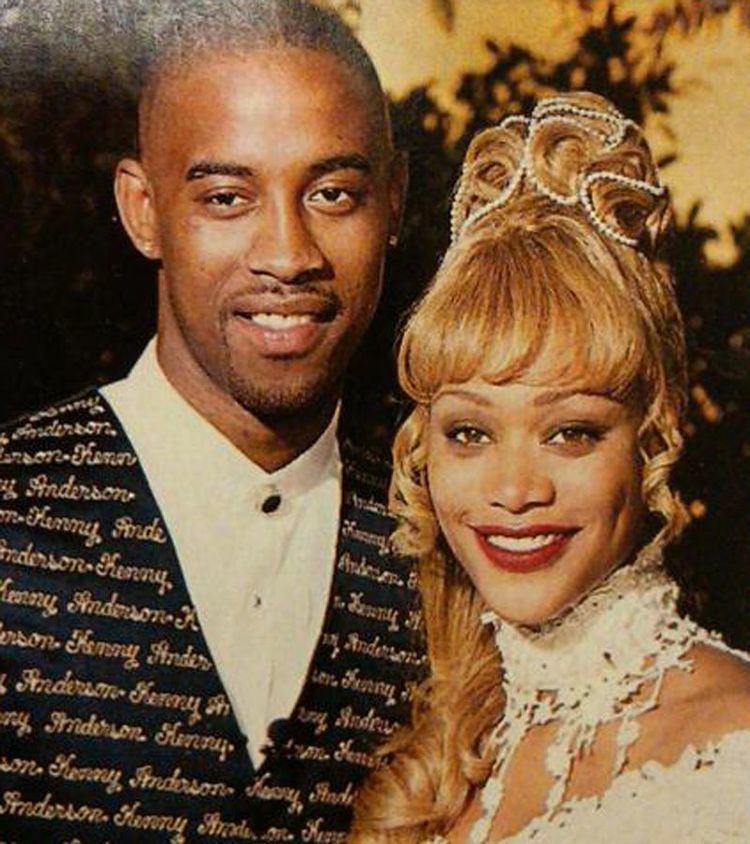 Kenny Anderson wearing a suit with his name on it and his wife wearing a white gown with red lipstick and blonde hair.