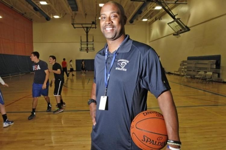 Kenny Anderson inside a basketball court wearing a gray shirt, a black watch with a ball in his hand with some basketball players on his back.