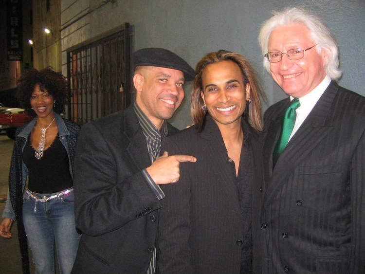 Kerry Gordy smiling with Reggie Benjamin and Tom Mesereau while he is wearing a black coat and black and gray striped long sleeves, and hat