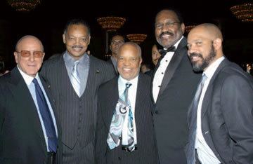 Kerry Gordy, on the right, smiling with the four men beside him and he is wearing a black coat, white long sleeves, and gray necktie