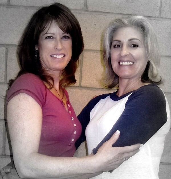 Kerry Lyn Dalton and her sister, Victoria Thorpe, smiling while hugging each other with a brick wall in the background. Kerry Lyn has shoulder-length blonde hair with a ring on her right finger wearing a white and black 3/4 sleeves baseball t-shirt. And Victoria has medium brown hair, a ring on her left finger, and a necklace while wearing a V-neck white-buttoned blouse.