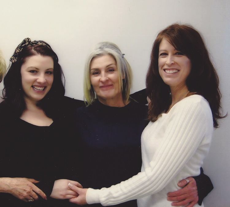 Christianna (Kerry's youngest child), Kerry Lyn, and Victoria are smiling while embracing each other on Christmas Day in 2011 (from left to right). Christianna has long wavy hair with a printed hairband on, and someone's hand with a finger ring holding her right wrist, is wearing a black long sleeve sweatshirt. Kerry Lyn has shoulder-length blonde hair, clipped on the left side, a silver finger ring on her right thumb, wearing a black long sleeve sweatshirt, while Victoria holding Christianna's right hand has long wavy brown hair, wearing a white knitted long sleeve sweatshirt.