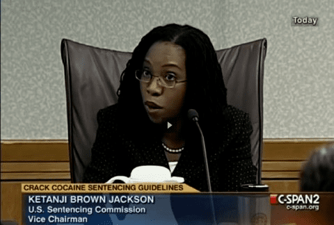 Ketanji Brown Jackson while talking during a hearing in the court with a microphone in front of her, sitting on a brown chair, with black hair, wearing eyeglasses, a pearl necklace, and a black blazer over a white top.