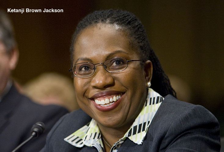 Ketanji Brown Jackson with a smiling face, black hair, wearing eyeglasses, earrings, and a gray blazer over a multi-colored polo shirt.