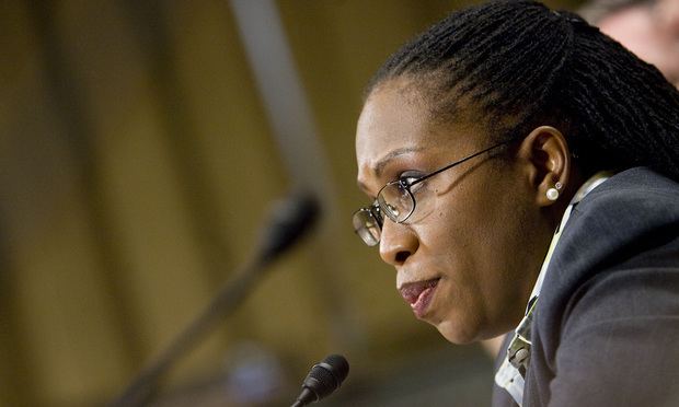 Ketanji Brown Jackson with a serious face with a microphone in front of her, with black hair, wearing eyeglasses, pearl earrings, and a gray blazer over a multi-colored polo shirt.