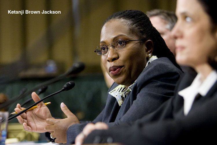 Ketanji Brown Jackson talking in front of a microphone while sitting beside her co-jurists and holding a pencil, with black hair, wearing eyeglasses, and a gray blazer over a multi-colored polo shirt.