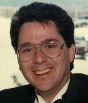 A younger Kevin Cosgrove smiling while wearing eyeglasses and a black suit.