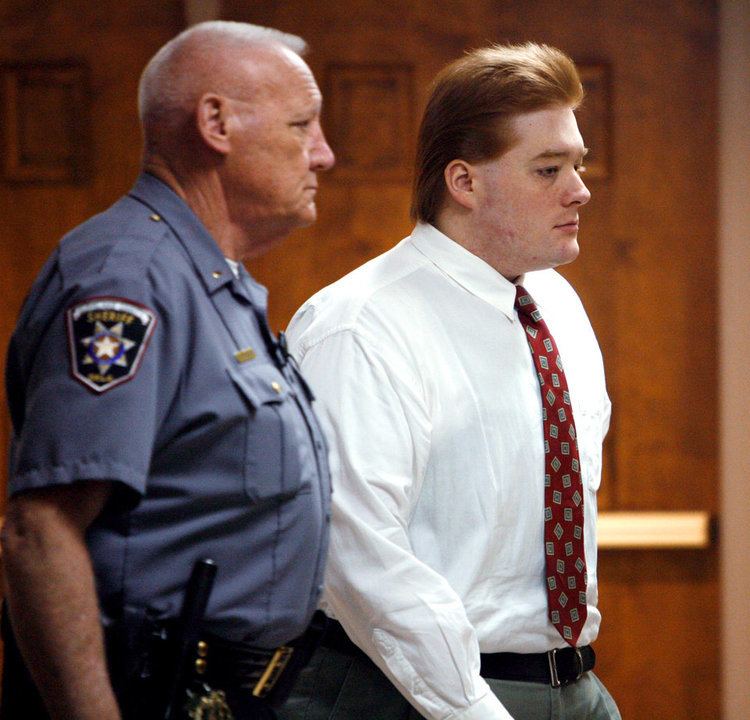 Kevin Ray Underwood and a sheriff officer with serious faces inside the court. Kevin with blonde hair, wearing white long sleeves, a red tie, a black belt, and gray pants while the sheriff officer is wearing a blue uniform and black pants.