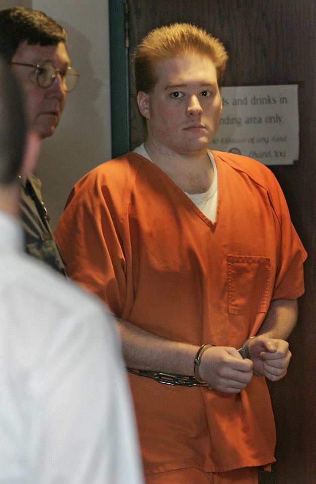 Kevin Ray Underwood with a serious face while looking at the man wearing white long sleeves and beside a man with a serious face and wearing eyeglasses. Kevin with blonde hair with a handcuff on his hands and wearing an orange prison shirt over a white shirt.