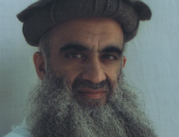 Khalid Sheikh Mohammed with a tight-lipped smile, mustache, and long beard while wearing a white shirt and brown hat