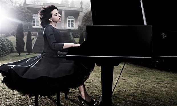 Khatia Buniatishvili playing a piano in an open field outside a mansion and wearing a black dress.