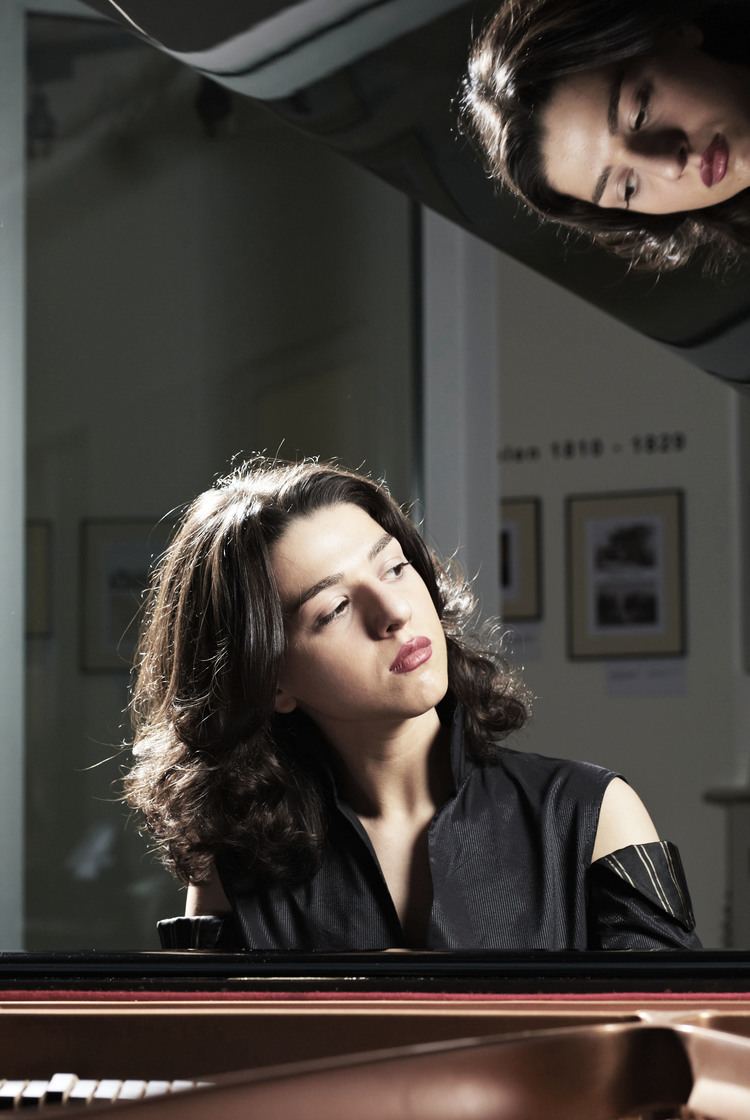 Khatia Buniatishvili posing sideways in a piano and wearing a black dress.