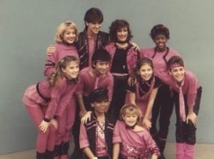 The cast of the children's television program, Kids Incorporated, smiling all together while wearing a black and pink costume