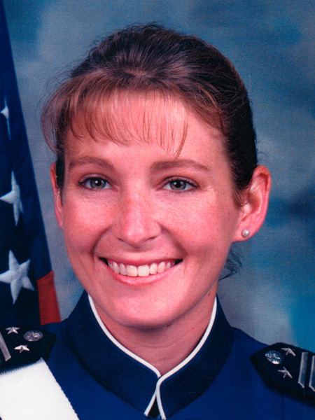 In front of a blue cloudy backdrop with USA flag at the right, Kim Campbell is smiling, she has brown hair, wearing a small pearl earrings, and a blue military uniform with black patch along with their ranks as military personnel.