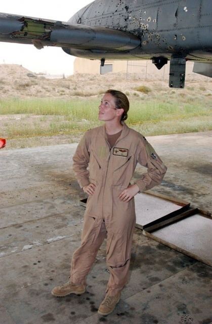 Outside a military base with a grass plains at the back with cream walls barracks, Kim Campbell is smiling, standing under the tail of a plane, both of her hands on waist, she has brown hair wearing a brown shirt under a US AIR FORCE flight suit with a patch on her left chest of a black bird and a brown boots,