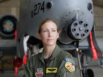 In front of a gray fighter jet with a round machine gun in front and 274 number painted, Kim Campbell is serious, standing, she has brown hair wearing a black shirt under an air force uniform. The uniform has three patches on the left a red wings  on a blue patch, 2nd from the left a patch of yellow with a black bird, at the right is a patch with a number A-10 over a yellow patch with a red line with white bird.