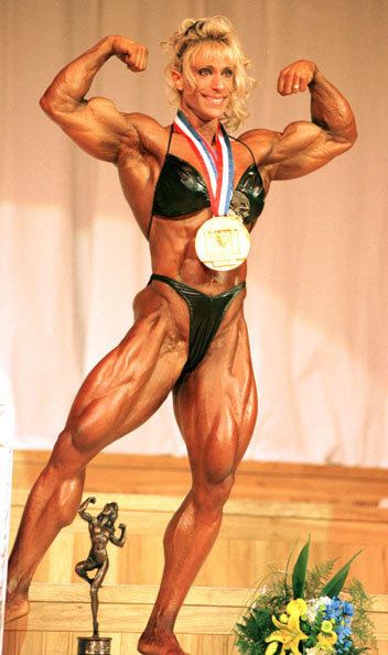 Kim Chizevsky-Nicholls smiling and flexing her muscles on stage and wearing a medal that she won as well as black bikini.