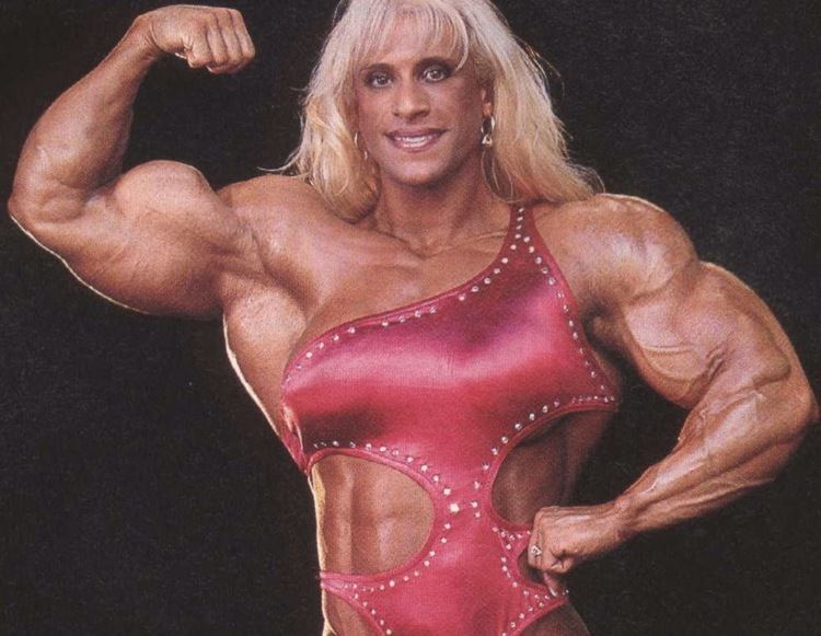 Kim Chizevsky-Nicholls smiling and flexing her muscles while wearing a pink bodybuilders outfit.