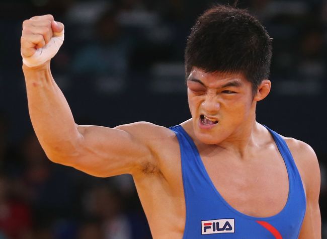 Kim Hyeon woo with a serious face raising his right arm in a closed fist is a male wrestler from South Korea with a bruise on his right eye wearing a blue FILA one-piece singlet.