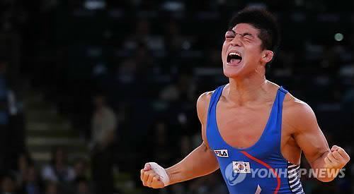 Kim Hyeon woo shouting with closed fists, a male wrestler from South Korea. With a crowd in the background, Kim celebrating his victory at the Greco-Roman 66-kilogram at the 2012 London Olympics with a bruise on his right eye, has black hair, a bandage on his right hand, and wearing a blue FILA with a South Korean flag one-piece singlet.