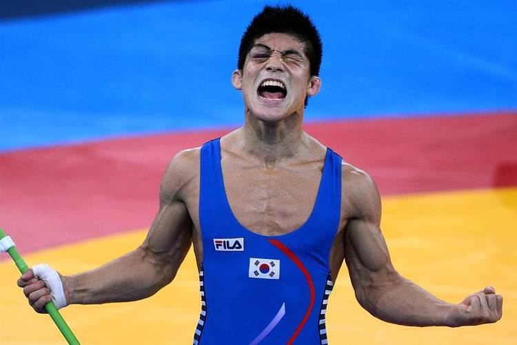 Kim Hyeon woo shouting with eyes closed while holding a green rod with a hand bandaged, a male wrestler from South Korea in the 2012 London Olympic Games; has bruised eye on the right, wearing a blue FILA with a small South Korean flag one-piece singlet.