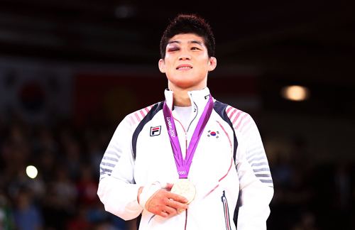 Kim Hyeon woo smiling while holding his gold medal on a violet strap, is a male wrestler from South Korea with a bruised eye, black hair, and a bandage on his right hand while wearing a white FILA with a South Korean flag Olympic jacket.
