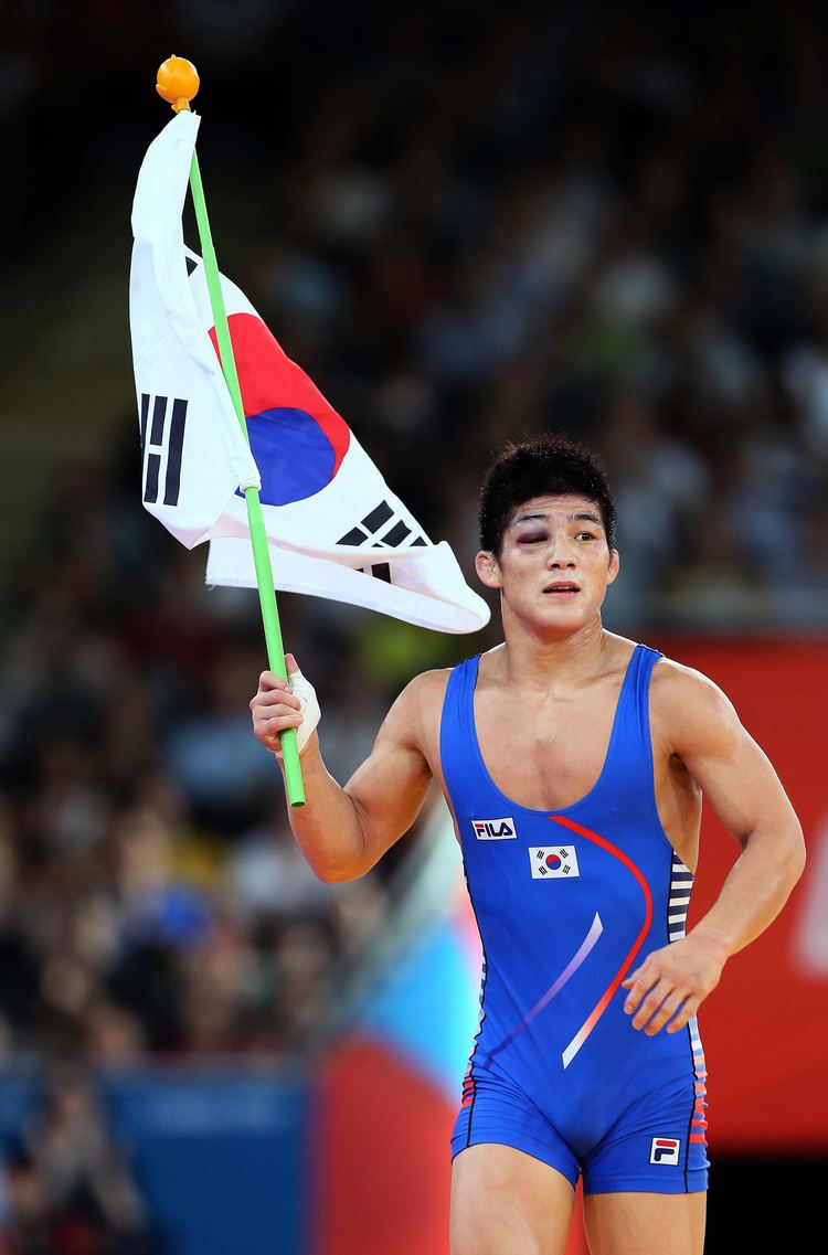 Kim Hyeon woo running while holding the Korean flag and looking at the side, a male wrestler from South Korea during the 2012 London Olympic Games with blurred crowds in the background. With his bruised right eye, and black hair, he is wearing a blue FILA with a small South Korean flag one-piece singlet.
