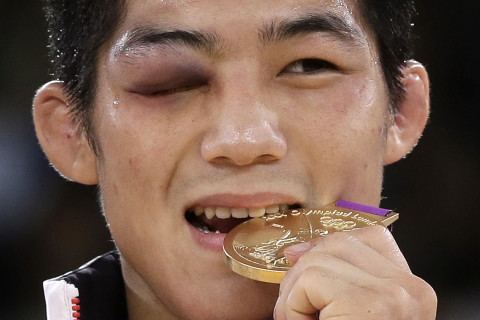 Kim Hyeon woo smiling while biting his gold medal, a male wrestler from South Korea, during the 2012 London Olympic Games with a bruised right eye, has black hair, wearing a white Olympic jacket.
