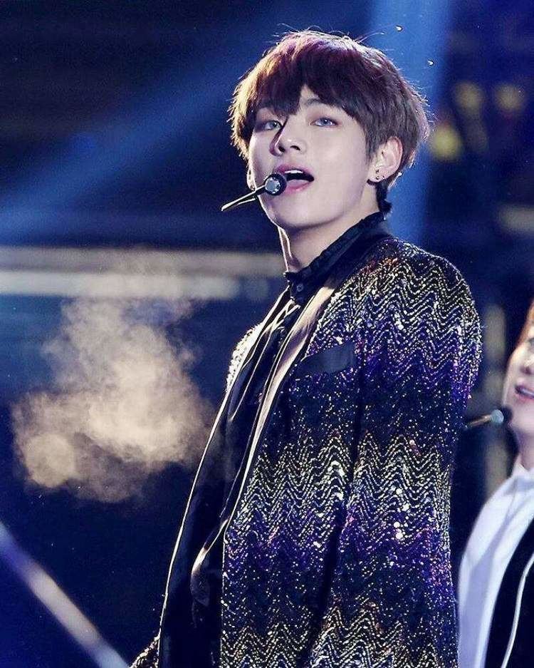 Kim Taehyung performing on stage, with a wired microphone, and wearing a fancy coat and black long sleeves.