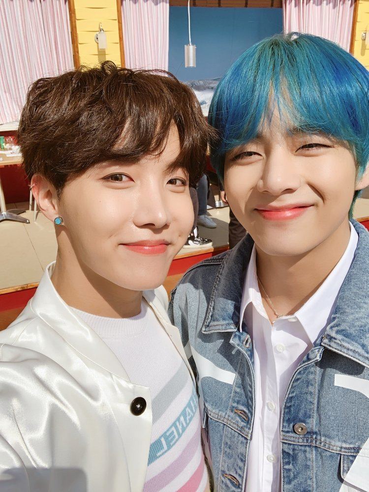 Kim Taehyung and J-Hope smiling, Taehyung with color blue hair, wearing a white polo shirt, and denim jacket while J-Hope wearing earrings and a white coat.