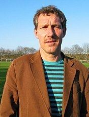 Kim Vilfort on a football field with a brown coat and a striped blue green shirt underneath.
