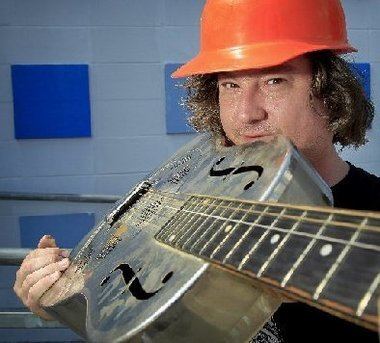 King Louie smiling while holding a silver guitar, with dark blonde wavy hair, and wearing an orange helmet and a black t-shirt