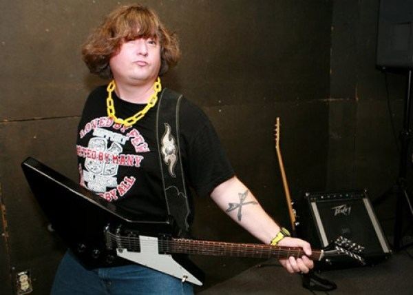King Louie Bankston holding a Gibson Explorer guitar, with a fierce look, brown short wavy hair, and a tattoo on his left arm  with a Peavey amplifier & Guitar in his background while wearing a black and yellow wristwatch, a yellow chain necklace, a black t-shirt with red and white prints, and blue pants