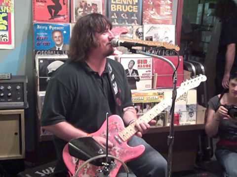 King Louie Bankston singing and playing the pink guitar while sitting on the chair with a microphone in front of him, magazines, and a girl holding a cam in the background. King Louie with a shoulder-length wavy hair is wearing a black polo shirt and gray pants