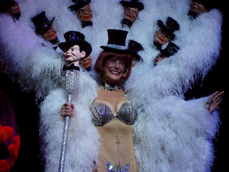 Kit Smythe smiling while wearing a black hat, silver bikini, and white wings