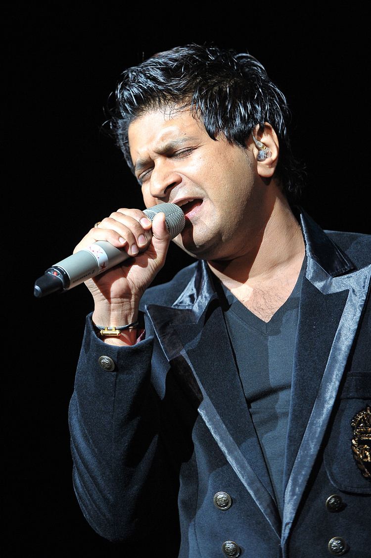 KK singing with closed eyes look while holding a microphone, he had black hair, wearing an earpiece, a bracelet, a ring on his right hand finger, and a black V-neck shirt under a black coat