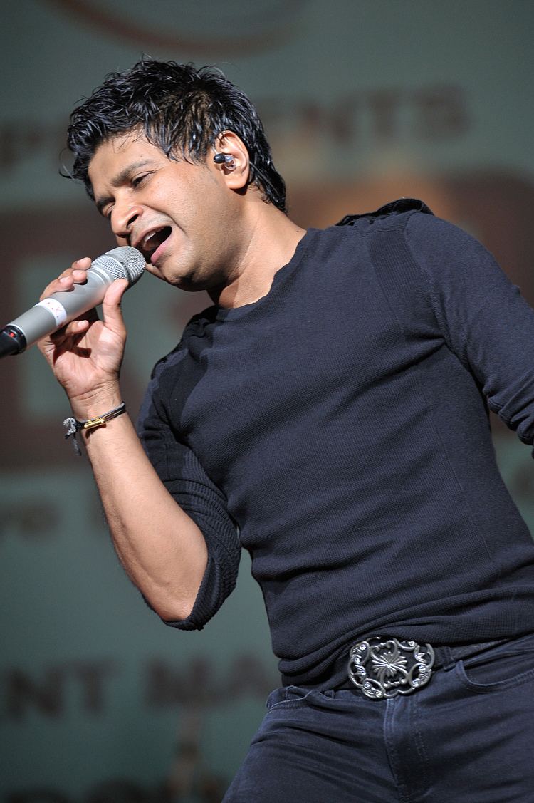 KK singing while holding a microphone, he had black hair, wearing an earpiece, bracelet, and a metal buckle black belt in denim jeans and a black longsleeve