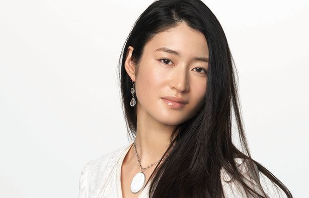 Koyuki Kato is serious in a gray background, has long black hair, wearing silver teardrop earrings, silver necklace with large white pendant, and a white top.