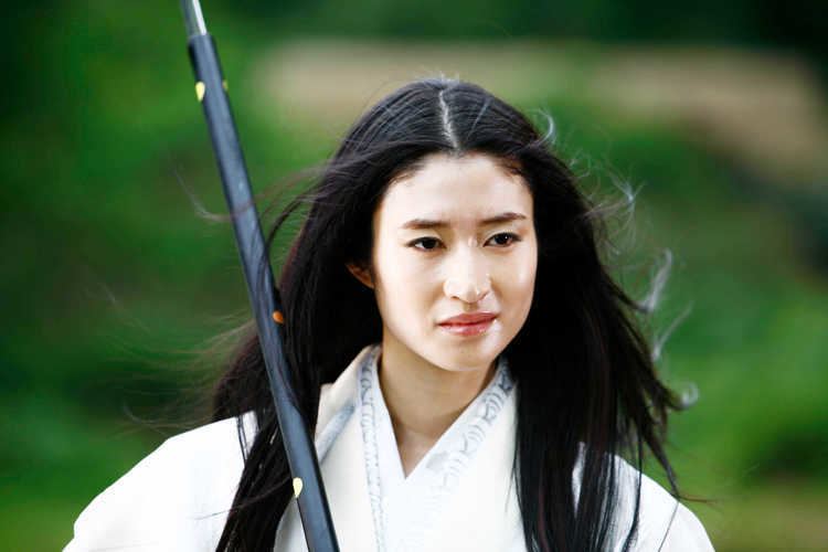 Koyuki Kato is serious, standing, holding a bō (black stick) on her right shoulder, has long black hair, and is wearing a all white kimono.