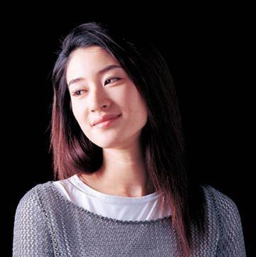 Koyuki Kato is smiling, standing looking at her right in a black background, has long black hair, wearing a white shirt under a gray see through top.