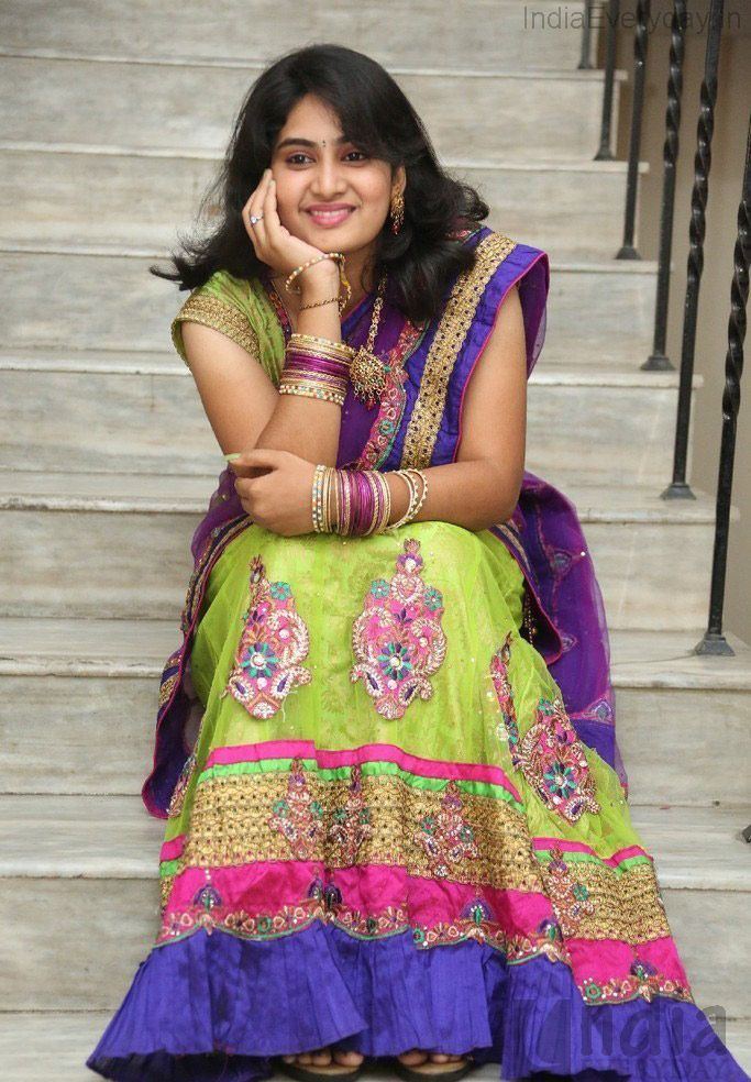 Krishnaveni smiling and sitting on the stairs while hand on her chin, with black wavy hair, bindi on her forehead, and wearing earrings, a necklace, bangles, and a colorful saree with a belt