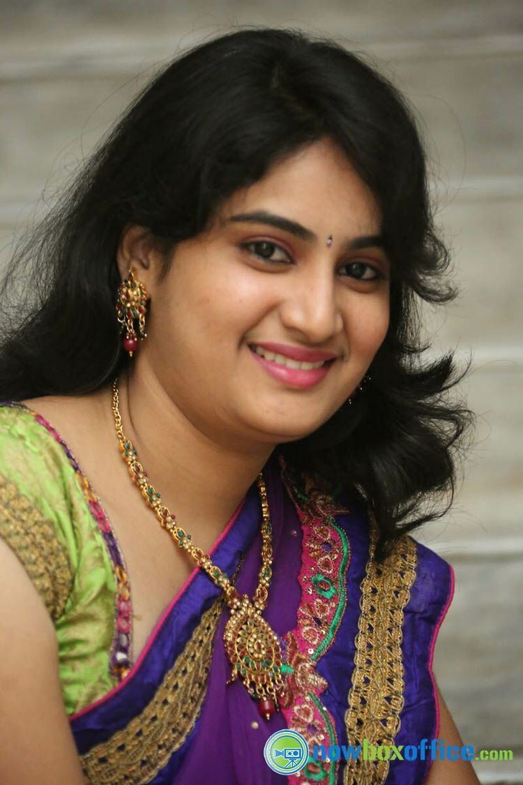 Krishnaveni smiling with black wavy hair and bindi on her forehead while wearing earrings, necklace, and a colorful saree
