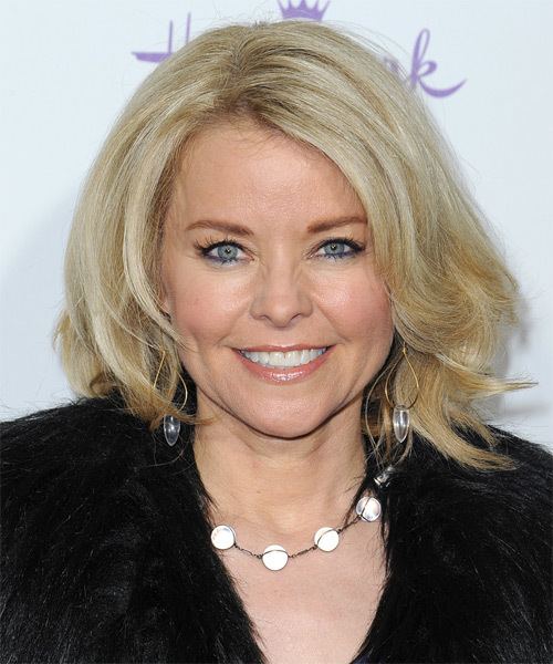 Kristina Wagner is smiling, has wavy blond hair, has blue eyes, wearing gold-silver earrings, a black necklace with a silver circle design, and a black fur coat.