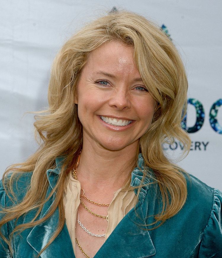 Kristina Wagner is smiling, has wavy blonde hair, and blue eyes, wearing a gold-silver necklace, and a flesh top under a blue coat.