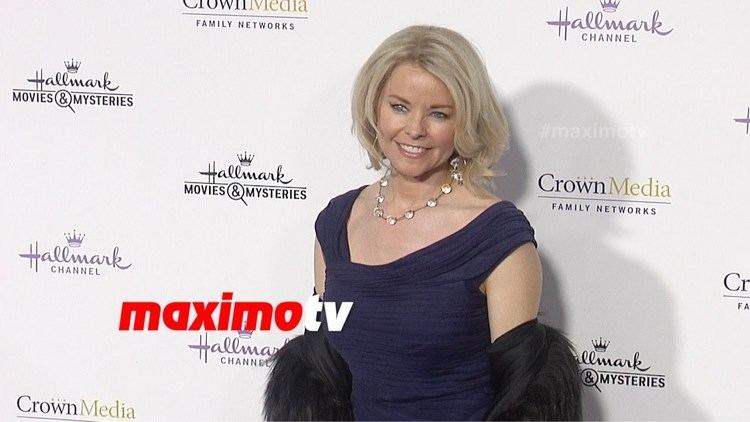 Kristina Wagner is smiling, has wavy blonde hair, wearing a silver earring on her right ear, a black necklace with white circles, a dark blue dress, and a black fur coat at the Hallmark TCA Party.