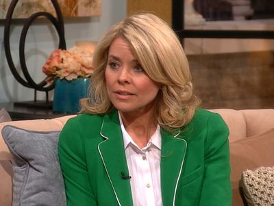 Kristina Wagner is serious, has wavy blonde hair with a microphone on her suit, wearing a white polo under a green suit, sitting on a brown sofa with a gray pillow on her (right) and 2 brown pillows on her (left).